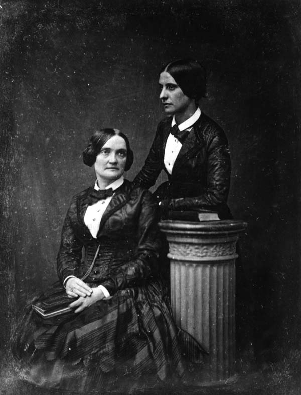 Two women posed for a portrait. One seated, one standing. 