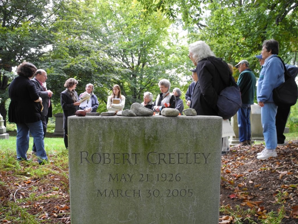 A gravestone reading "Robert Creeley" in the foreground with a tour group of people gathered in the background.