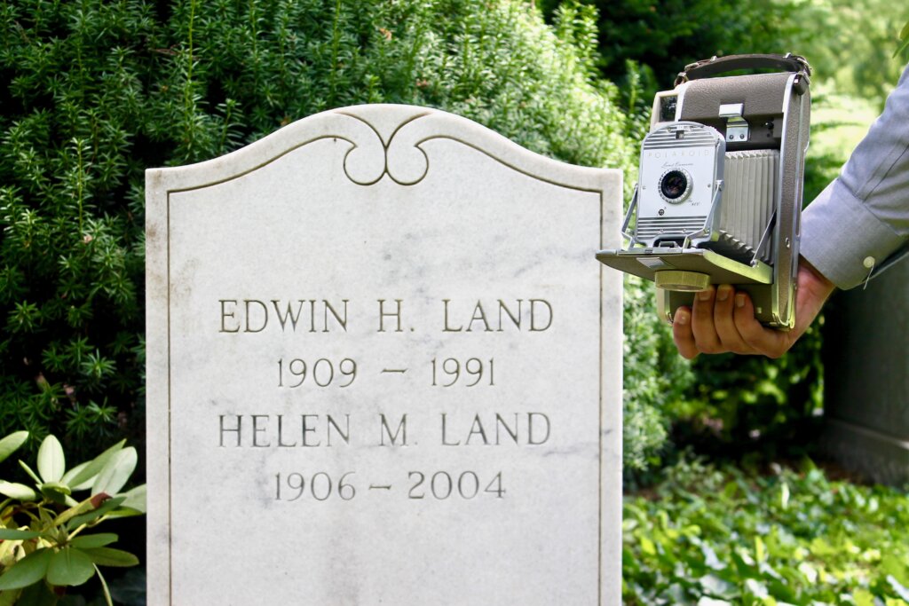 a marble gravestone with a vintage polaroid camera held in front of it. 
