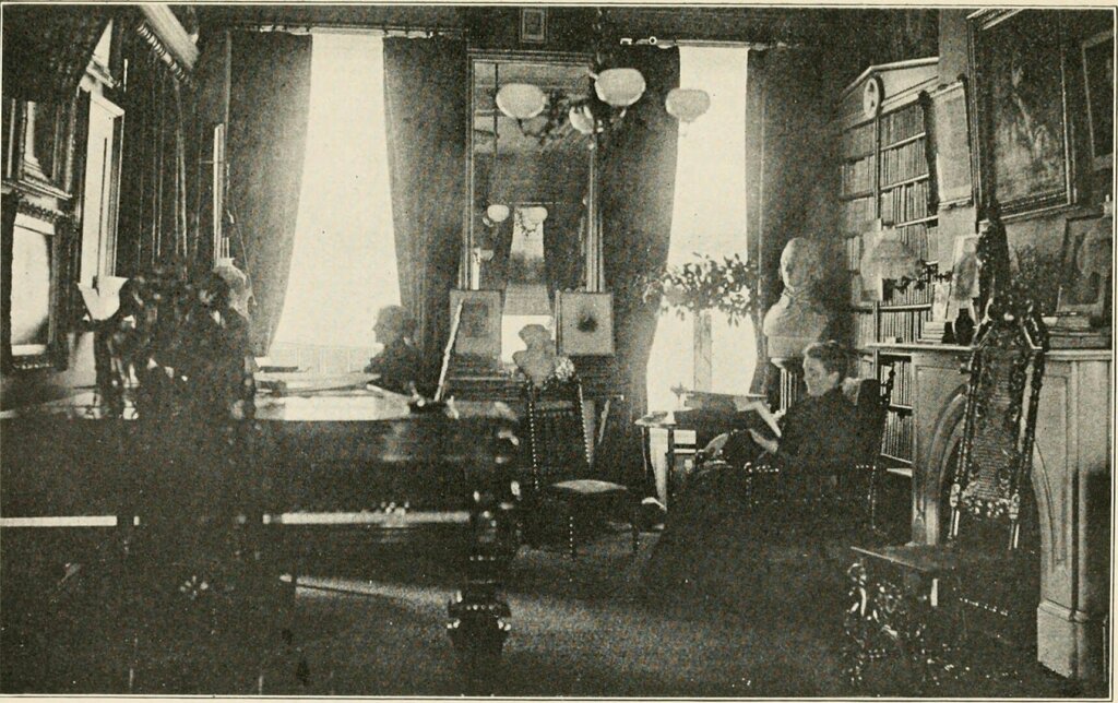 A photograph of a room where one woman sits by the fireplace and another sits by the window. 