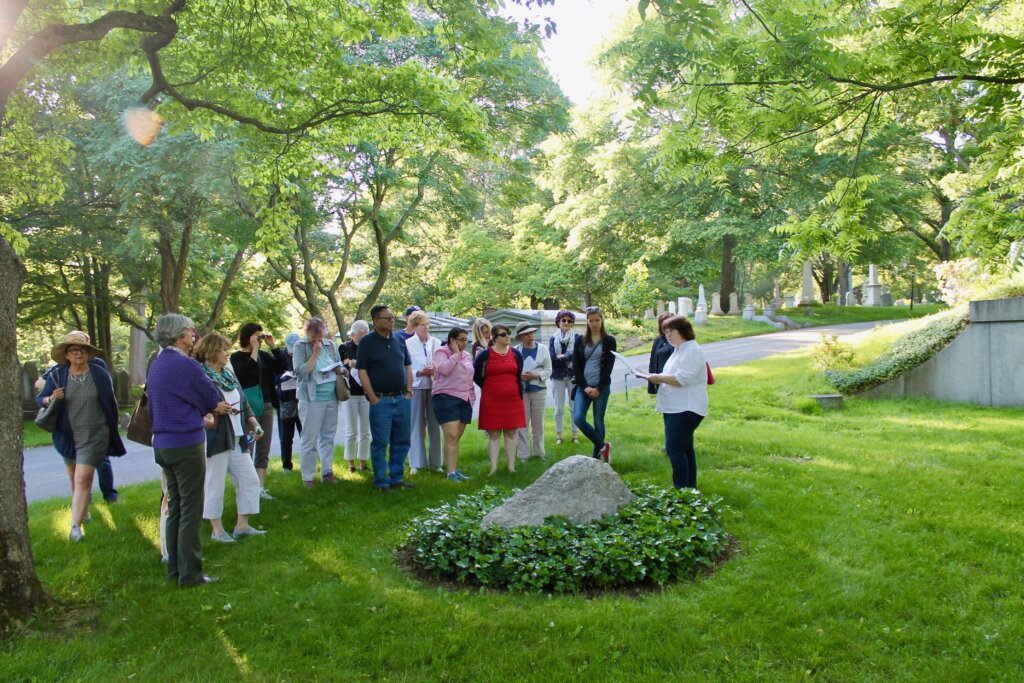 A tour group gathers around a stone grave surrounded by ivy