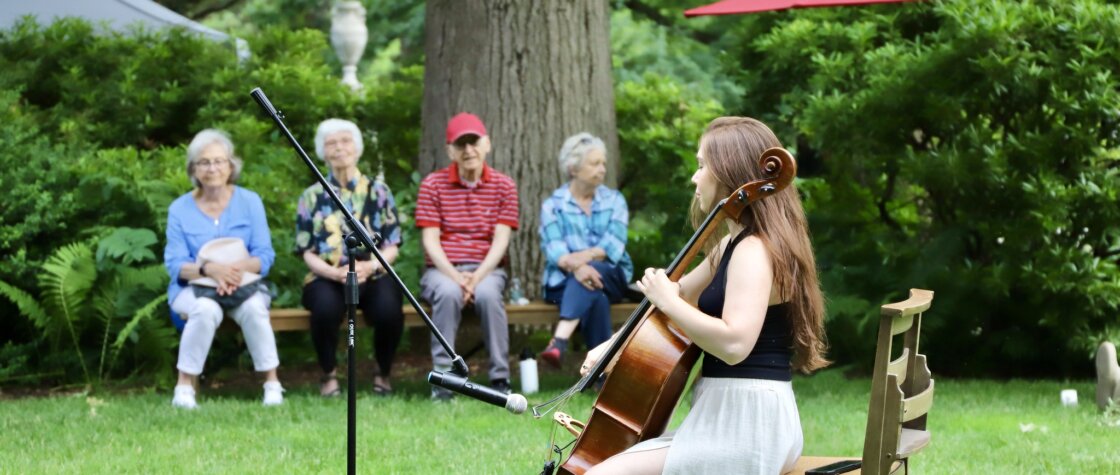 a seated woman plays a cello outdoors in front of a small audience