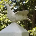 A marble dove with spreading wings perches on the top of an obelisk. The marble is deteriorated but the shape of the bird is still evident.