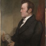 Portrait painting of Johann Gaspar Spurzheim. He is painted in profile and points to the skull of a bust he holds in his lap.